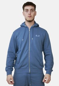 Felpa blu con zip realizzata in tessuto morbido, dotata di zip frontale in metallo, cappuccio regolabile con coulisse e un piccolo logo sul petto.