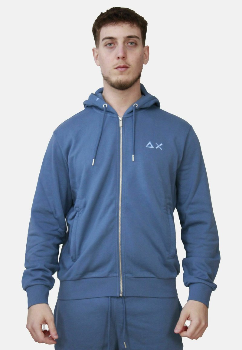 Felpa blu con zip realizzata in tessuto morbido, dotata di zip frontale in metallo, cappuccio regolabile con coulisse e un piccolo logo sul petto.