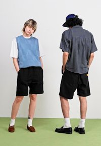 Zwei Models tragen legere Kleidung; eines trägt eine blaue Patchwork-Weste und schwarze Shorts, das andere ein kariertes Hemd und schwarze Shorts.