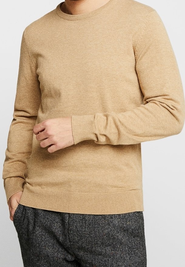 Pull en tricot beige avec un col rond, des poignets et un ourlet côtelés. Coupe ajustée et texture douce, présenté avec un pantalon gris à motifs.
