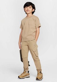 Μπεζ μπλουζάκι Nike με τσέπη, συνδυασμένο με αντίστοιχο παντελόνι cargo με πλαϊνές τσέπες και μεταλλικά χρυσά αθλητικά παπούτσια. Ουδέτερο χρωματικό συνδυασμό.