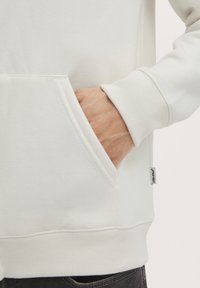 Hvit sweatshirts med frontlomme, ribbestrikkede cuffs og midjebånd, samt en liten merkeetikett på siden. Myk stofftekstur synlig.