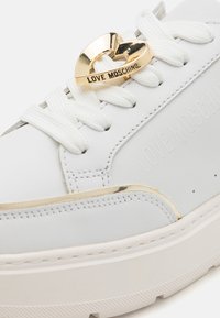 White leather sneaker with gold heart-shaped hardware and embossed "LOVE MOSCHINO" branding. Features white laces and textured outsole.