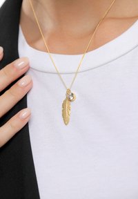 Gouden ketting met een hangertje in de vorm van een veer met gedetailleerd vakmanschap en een kleine ronde steen als accent. Draagt op een witte top.