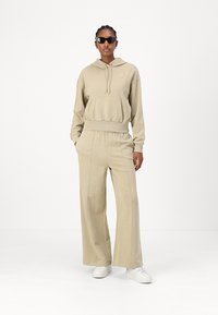 Beige hoodie med snörning och framficka, matchad med vida beige byxor. Buren med vita sneakers och svarta solglasögon.