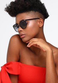 Sunglasses with black round frames, dark lenses, and gold accents. The model wears a red off-shoulder top and rests her hand on her chin.