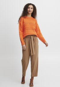 Pull en maille orange avec un motif, associé à un pantalon brown taille haute avec une ceinture nouée et des chaussures à talons marron.