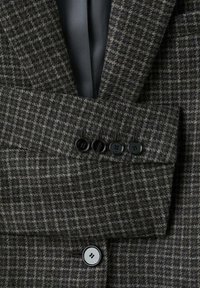 Marynarka w szary wzór houndstooth z czarnymi guzikami, z wywijanym kołnierzem i wyraźnymi ramionami. Teksturowany materiał, subtelny połysk.