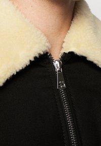 Czarna kurtka z zamkiem błyskawicznym z puszystym beżowym kołnierzem; ma metalowy zamek i teksturowany materiał; zbliżenie na kołnierz i obszar klatki piersiowej.