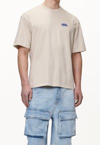 Beige t-shirt med kort ärm och en blå logotyp på bröstet, tillverkad av bomull, med en avslappnad passform och rundad halsringning, kombinerad med ljusa denim cargobyxor.
