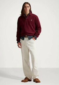 Bordowa koszula polo z długim rękawem, nałożona na niebieską i zieloną koszulę w kratę. Kremowe spodnie typu wide-leg oraz brązowe buty dopełniają całości.