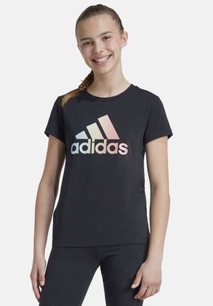 Camiseta de algodón negra con un gran logo de Adidas en degradado reflectante. Mangas cortas y cuello redondo. Modelo vistiendo leggings negros.
