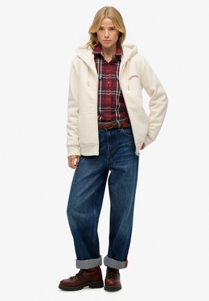 Kvinde iført en cremet lynlås hoodie over en rød rutig skjorte, løse opsmøgede blå jeans, brun bæltetaske og brune snørebåndsstøvler, stående foran en hvid baggrund.