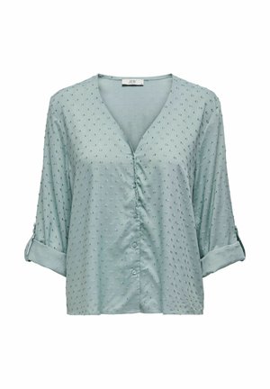 Blouse bleu clair à manches courtes avec un col en V, ornée de points texturés et de boutons sur le devant. Les manches retroussées ajoutent une touche au design.