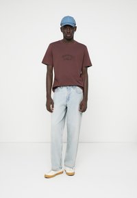 Jonge man die staat, gekleed in een bruin T-shirt, lichte blauwe jeans, witte sneakers en een lichtblauwe pet tegen een effen witte achtergrond.