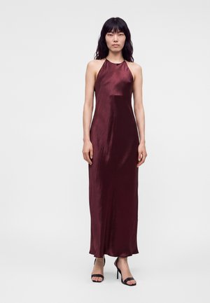 Femme portant une robe dos nu en satin rouge foncé avec des sandales à talons hauts noires, debout devant un fond clair uni.