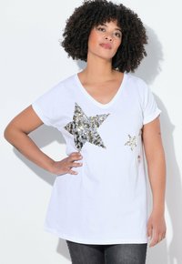 Bílý trikový top s V-výstřihem a velkými metalickými zlatými flitrovými hvězdami na přední straně, krátkými ohrnutými rukávy a volným střihem.