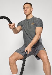 Grå treningsskjorte med teksturert stoff og oransje logo, kombinert med matchende shorts. Personen holder et tykt svart tau.