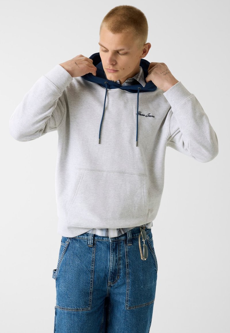 Jeune homme ajustant la capuche d'un sweat à capuche gris clair avec des cordons bleu marine, portant un jean bleu avec des lunettes accrochées à la poche.