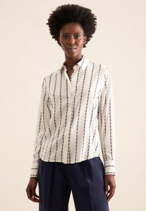Camicia bianca con motivo a catena verticale blu, colletto a bottoni e maniche lunghe. Presenta un taglio dritto e polsini con dettagli blu.