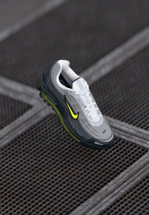 Szaro-biały sneaker Nike Max Air z neonowo-żółtymi akcentami, zawieszony nad metalową kratą podłogi.