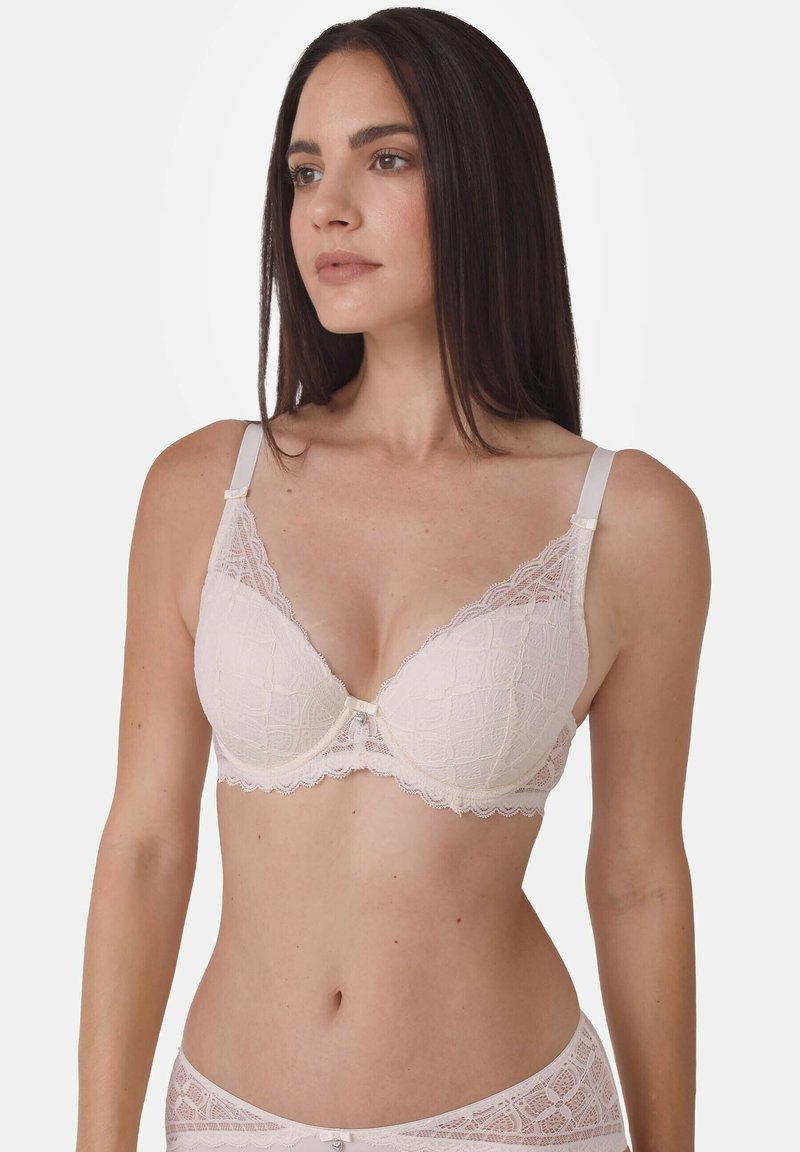 Frau trägt einen passenden hellrosa Spitzen-BH und Spitzen-Slip, steht mit entspannten Händen da und schaut seitlich vor einem weißen Hintergrund.