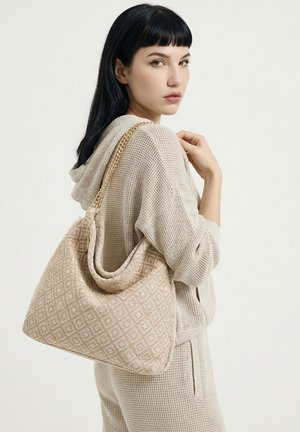 Jeune femme aux cheveux noirs portant un hoodie en maille beige et un pantalon, portant un sac à bandoulière beige à motifs avec une chaîne dorée.