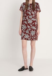 Robe à motifs floraux rouges avec des manches courtes et une coupe droite, accompagnée de chaussures noires à enfiler. Tissu lisse avec une coupe décontractée.