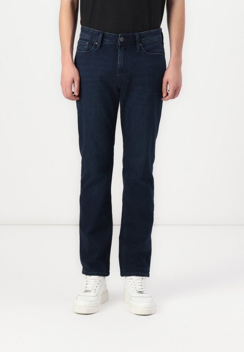 Jeans din denim albastru închis cu croială dreaptă, având buzunare frontale, bucle pentru curea și un finisaj texturat discret. Puși în completare cu adidași albi.