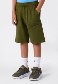Shorts en polaire vert avec une taille à cordon de serrage, dotés de deux poches latérales. Associés à des baskets grises et des chaussettes blanches à la cheville.
