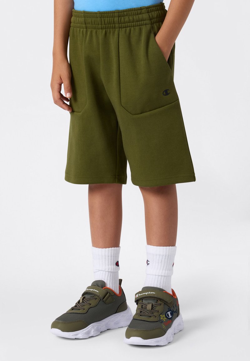 Shorts en polaire vert avec une taille à cordon de serrage, dotés de deux poches latérales. Associés à des baskets grises et des chaussettes blanches à la cheville.