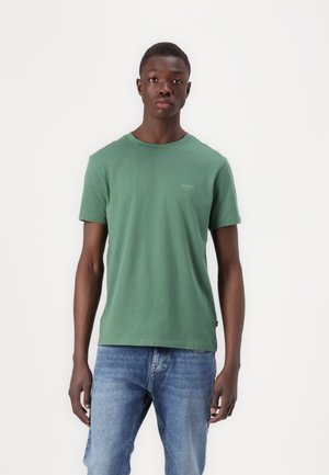 Green cotton t-shirt with a crew neck, short sleeves, and a small logo on the left chest. Worn with light blue denim jeans.