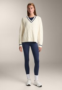 Cremfarbener Strickpullover mit Rautenmuster und Marineblauem Besatz, kombiniert mit Marineblauen Leggings und weißen Sneakers. Elastische Bündchen an den Ärmeln.
