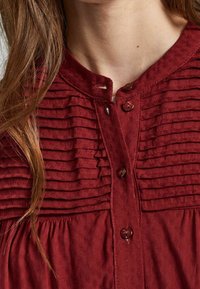 Burgundy blus med plisserad frontdetalj och rund halsringning, försedd med knappar och ett texturerat tyg. Inga synliga mönster eller grafiker.