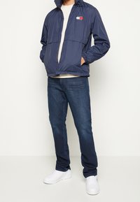Chaqueta cortavientos de color navy con puños y cintura elásticos, con un logo en el pecho; combinada con unos jeans azul oscuro y zapatillas blancas.