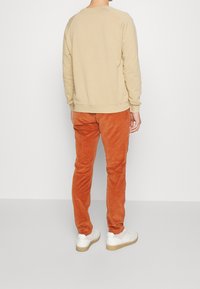 Persona vestida con sudadera beige, pantalones de pana anaranjados quemados y zapatillas blancas, de pie frente a una pared blanca sencilla.
