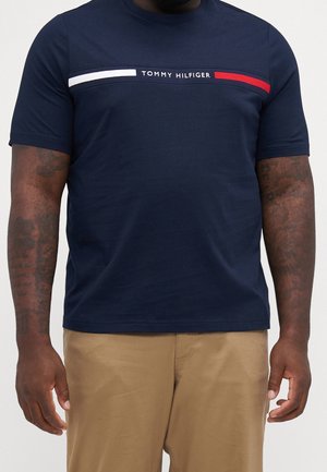 Uomo che indossa una maglietta Tommy Hilfiger blu navy con strisce bianche e rosse sul petto e pantaloni beige, tatuaggi visibili sulle braccia.