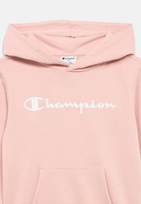 Sweat à capuche rose clair en tissu doux, avec une poche kangourou à l'avant et un grand logo blanc "Champion" sur la poitrine.