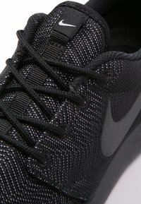 Svart idrottssko med en texturerad mesh-ovandel, platta svarta snören och en framträdande svart Nike-logotyp på sidan. Subtil lyster.