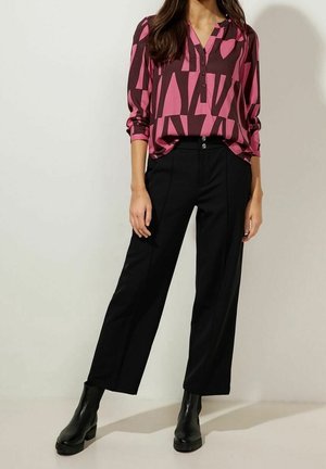 Vrouw draagt een roze en bruine blouse met geometrisch patroon, zwarte broek met wijde pijpen en zwarte enkellaarzen, staand op een lichte vloer.