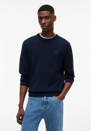 Ung mann iført en mørkeblå genser med rund hals over en hvit skjorte, kombinert med lyseblå jeans, stående med en hånd i lommen.