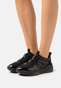 Scarpe da ginnastica nere realizzate con materiali lucidi e in rete, caratterizzate da lacci neri e accenti con logo, progettate con una suola spessa e una forma arrotondata.