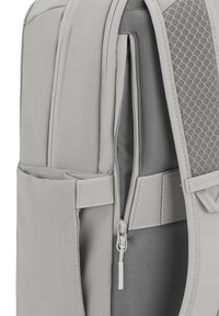 Grå ryggsäck med en slät, texturerad yta, ett nätpanel, flera fickor och en dragkedja för enkel åtkomst.