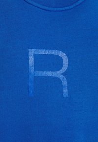 Blå bomulls t-shirt med ett något upphöjt, ljusblått bokstav "R" på framsidan. Tyget har en ribbad textur och en rundad halsdesign.