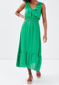 Robe maxi verte sans manches avec un col à volants, une taille smockée et un ourlet à niveaux. Fabriquée en tissu léger avec une texture fluide.