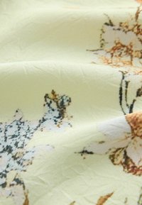 Light yellow fabric with a delicate floral pattern in muted brown, black, and white tones, softly folded with visible texture.