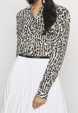 Kobieta ubrana w czarno-białą bluzkę w zwierzęcy wzór z długimi rękawami, zestawioną z białą plisowaną spódnicą i perłowymi kolczykami.