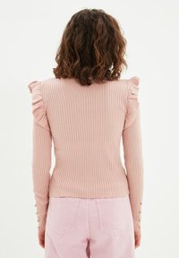 Maglietta a maniche lunghe di color rosa chiaro con finitura a coste, caratterizzata da dettagli arricciati sulle spalle e bottoni sui polsini. Texture liscia e design aderente.