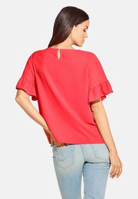 Rotes kurzärmeliges Oberteil mit Rüschensäumen und einem Schlüsselloch-Rückenverschluss, gefertigt aus einem glatten Stoff. Kombiniert mit hellblauen Jeans.