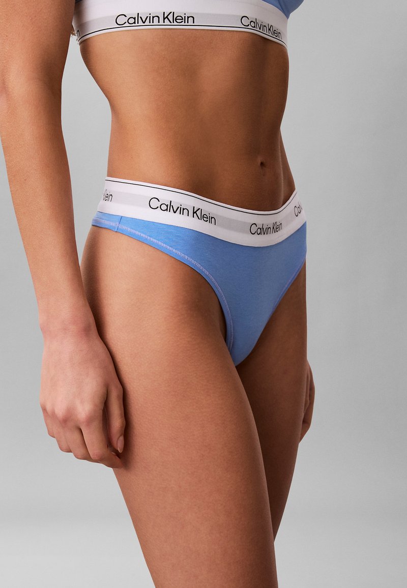 Close-up van een persoon die lichtblauwe Calvin Klein onderkleding en een bijpassende sportbeha draagt tegen een neutrale grijze achtergrond.